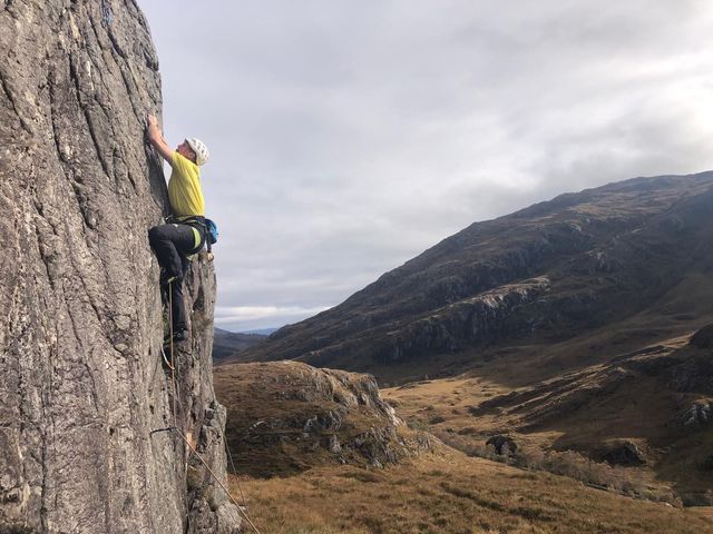 Dave rock climbing in a yellow T-shirt above a Highland glen